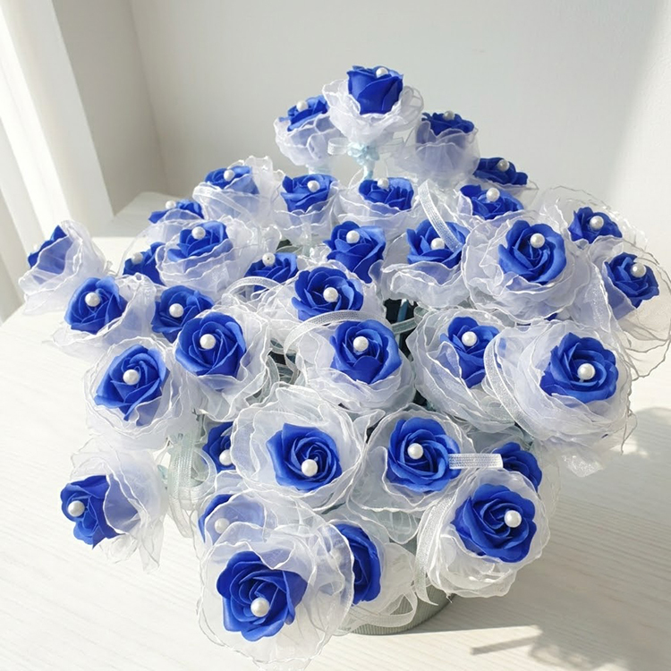 在明亮的室內陽光下，一整盆色彩鮮豔的深藍色玫瑰肥皂花，每朵花心都鑲嵌著優雅的珍珠並包裹著白色蕾絲花邊，精緻地擺放在簡約乾淨的白色木紋桌面上。