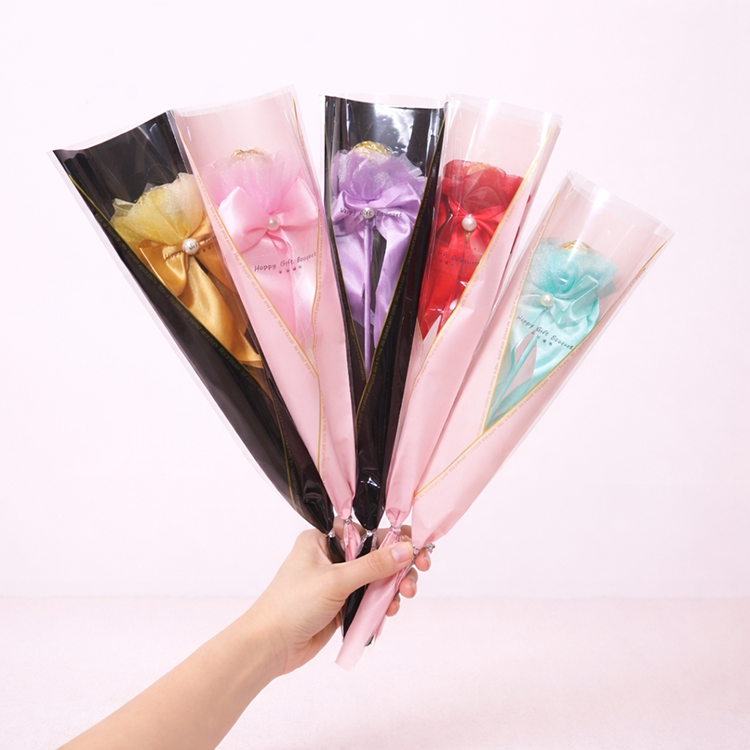 五色金莎花束展示圖,一隻手拿著金色、粉紅、薄荷綠、紅色與紫色單枝金莎棒棒花,搭配緞帶與珍珠裝飾,背景為柔和淺粉色 #FDEBED。適合母親節禮物、情人節禮物、畢業禮物、小禮物、金莎花束商品圖。