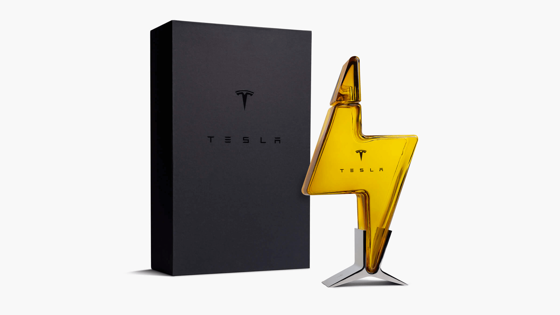 Tesla Tequila 限量龍舌蘭酒，手工吹製閃電造型琥珀色玻璃瓶，瓶身壓印 Tesla 標誌，立於銀色三腳金屬底座上。左側為霧面黑色精裝禮盒，同樣印有 Tesla Logo 與品牌字樣，整體呈現高端收藏品質感。