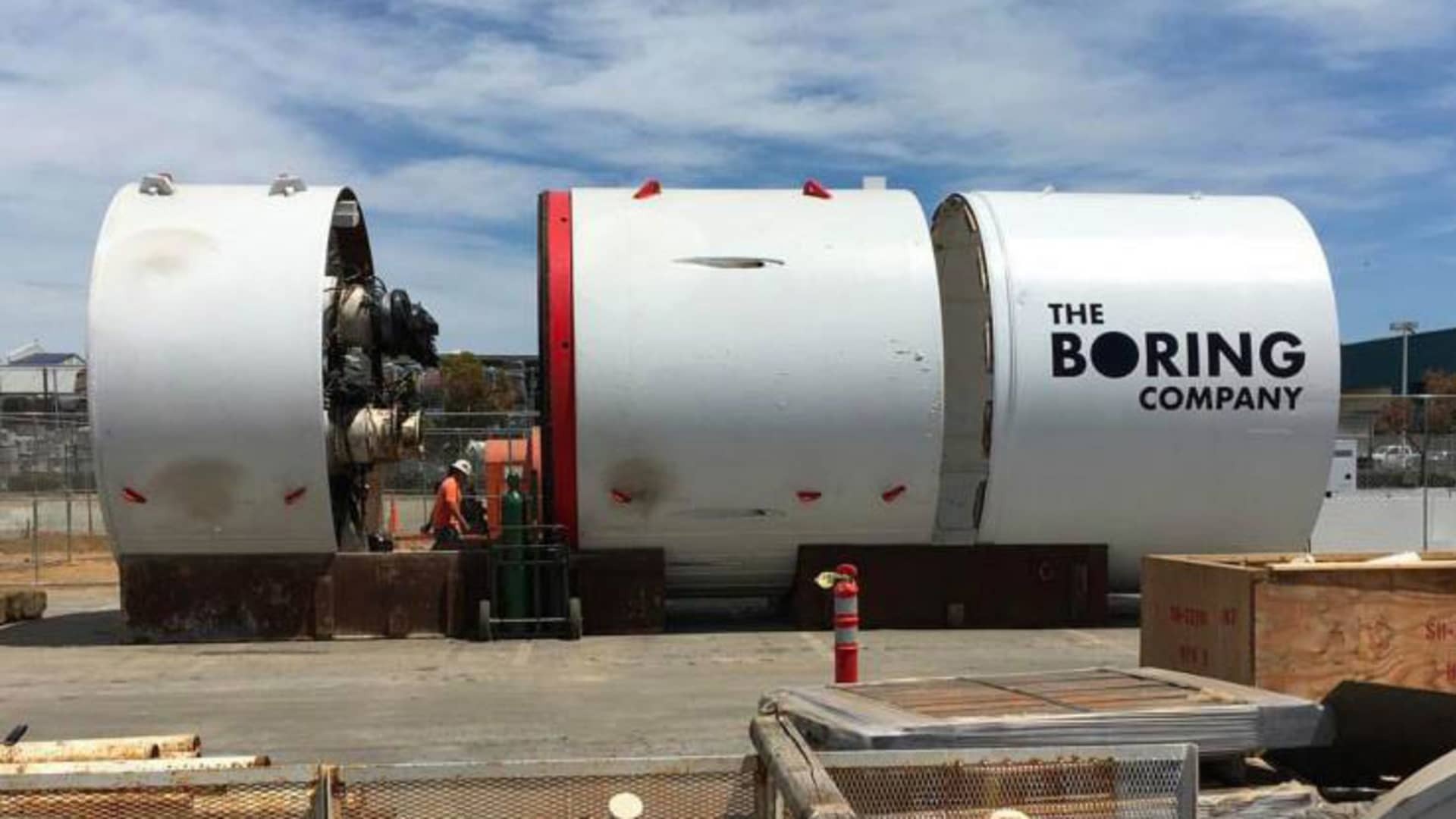 The Boring Company 巨型隧道鑽掘機實拍，白色圓柱形機身印有品牌名稱，工人站於機器入口旁進行作業，呈現設備的龐大體積。