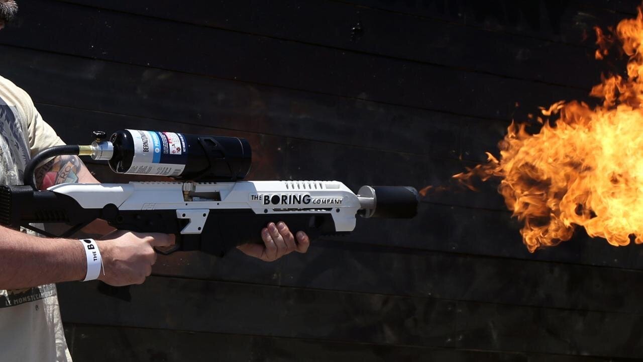 The Boring Company 限量「Not-a-Flamethrower」實拍，使用者手持白色槍身印有品牌字樣的裝置，槍口噴出大片火焰，上方裝有燃料罐。