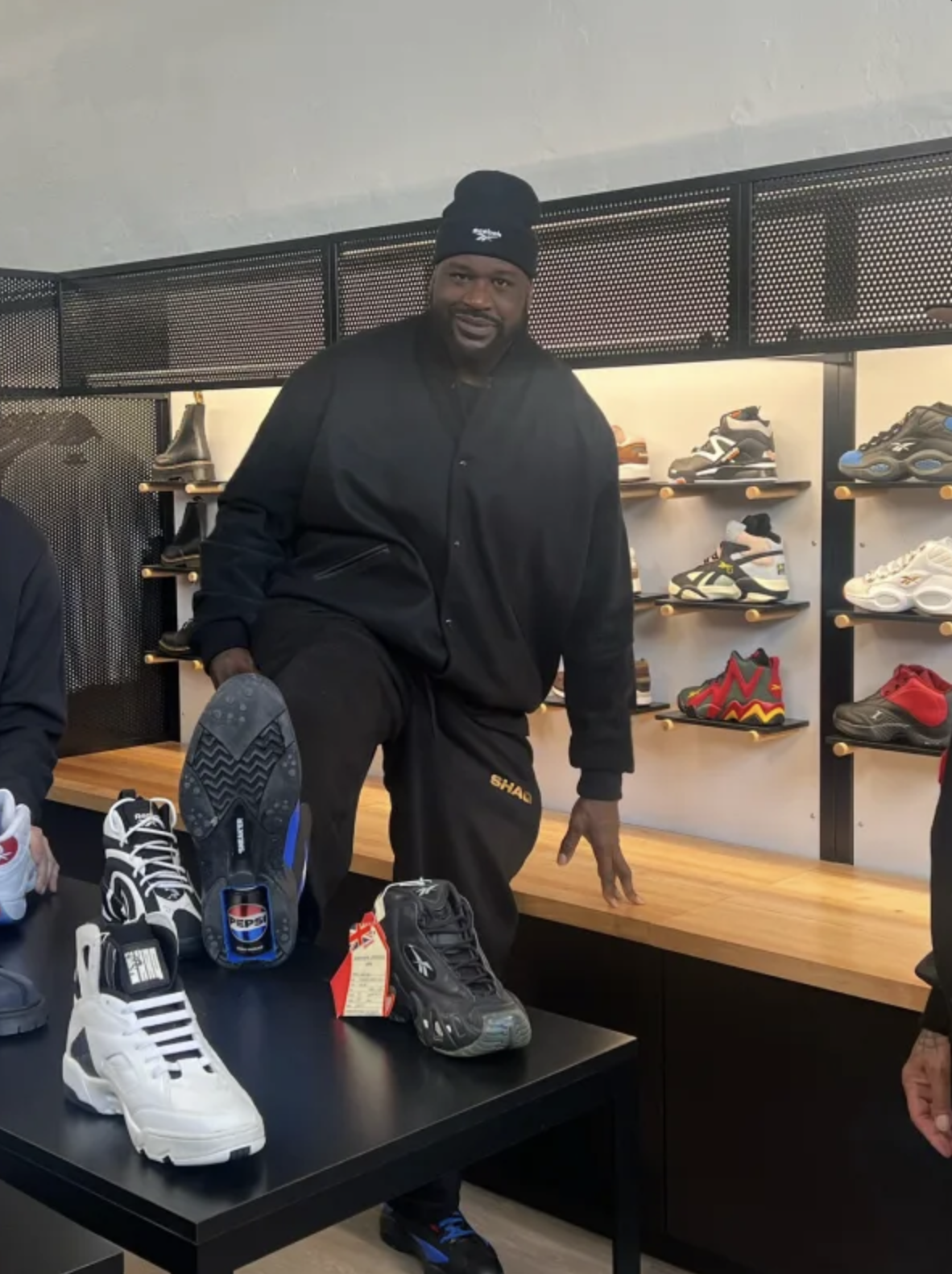 俠客歐尼爾（Shaquille O'Neal）於球鞋店內展示 Pepsi x Reebok 聯名限量鞋款，抬腳秀出鞋底內嵌的迷你百事可樂罐，桌上並排多款 Reebok Shaq 系列球鞋。