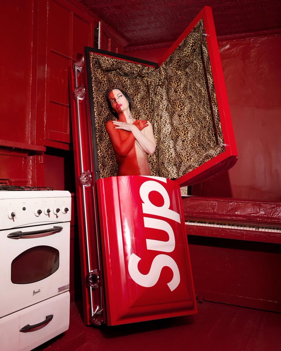 Supreme x Titan Casket 聯名棺材官方宣傳照，模特兒身著紅色緊身衣坐於開蓋棺材中，豹紋內裡清晰可見，置於全紅色調房間內，旁邊搭配白色烤箱與鋼琴，呈現品牌一貫的強烈視覺風格。