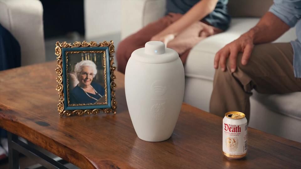 Eternal Playlist Urn 宣傳影片截圖，骨灰罈置於木質茶几上，左側擺放老奶奶遺照，右側放一罐 Liquid Death，背景為坐在沙發上的家人，呈現骨灰罈融入日常家居場景的使用情境。