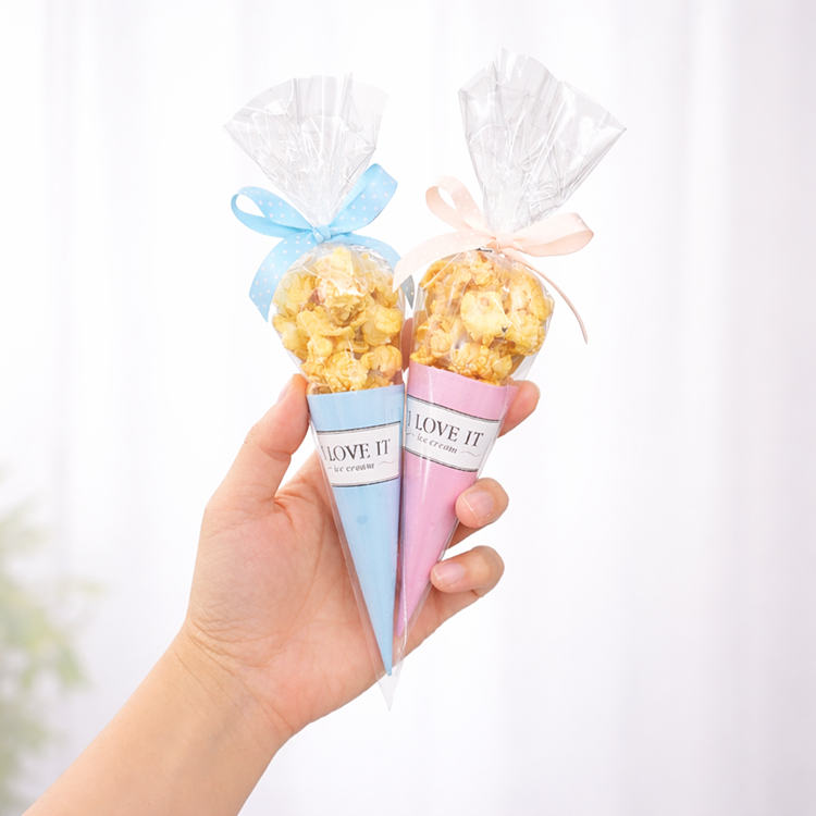 手持兩支甜筒造型爆米花禮物，一支為藍色包裝搭配藍色蝴蝶結，另一支為粉色包裝搭配粉色蝴蝶結，透明包裝內可見焦糖爆米花，背景明亮簡潔。