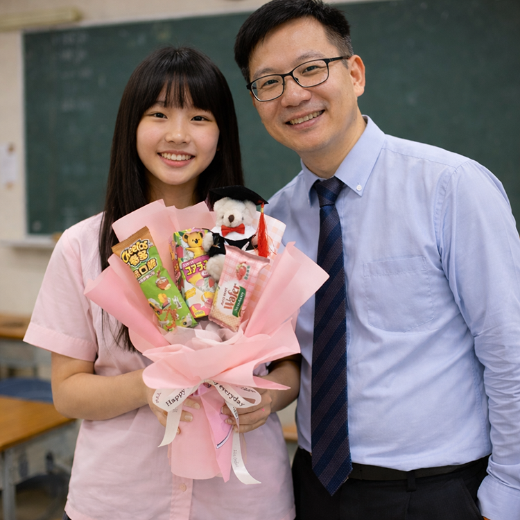 兩人在教室裡合影,其中學生手持粉色包裝紙包裹的畢業熊熊零食花束,花束裡餅乾、威化餅,以及一隻戴著畢業帽和學士服的熊熊,綁著寫有『Happy day』黑色字樣的白色緞帶,背景是黑板與課桌椅,呈現慶祝或畢業的氛圍。