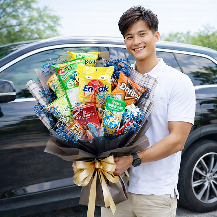 男生拿著一束裝滿各種知名品牌零食（如多力多滋、巧克力）的創意花束，採用黑色與格紋包裝紙及金色緞帶裝飾，背景為黑色車前面