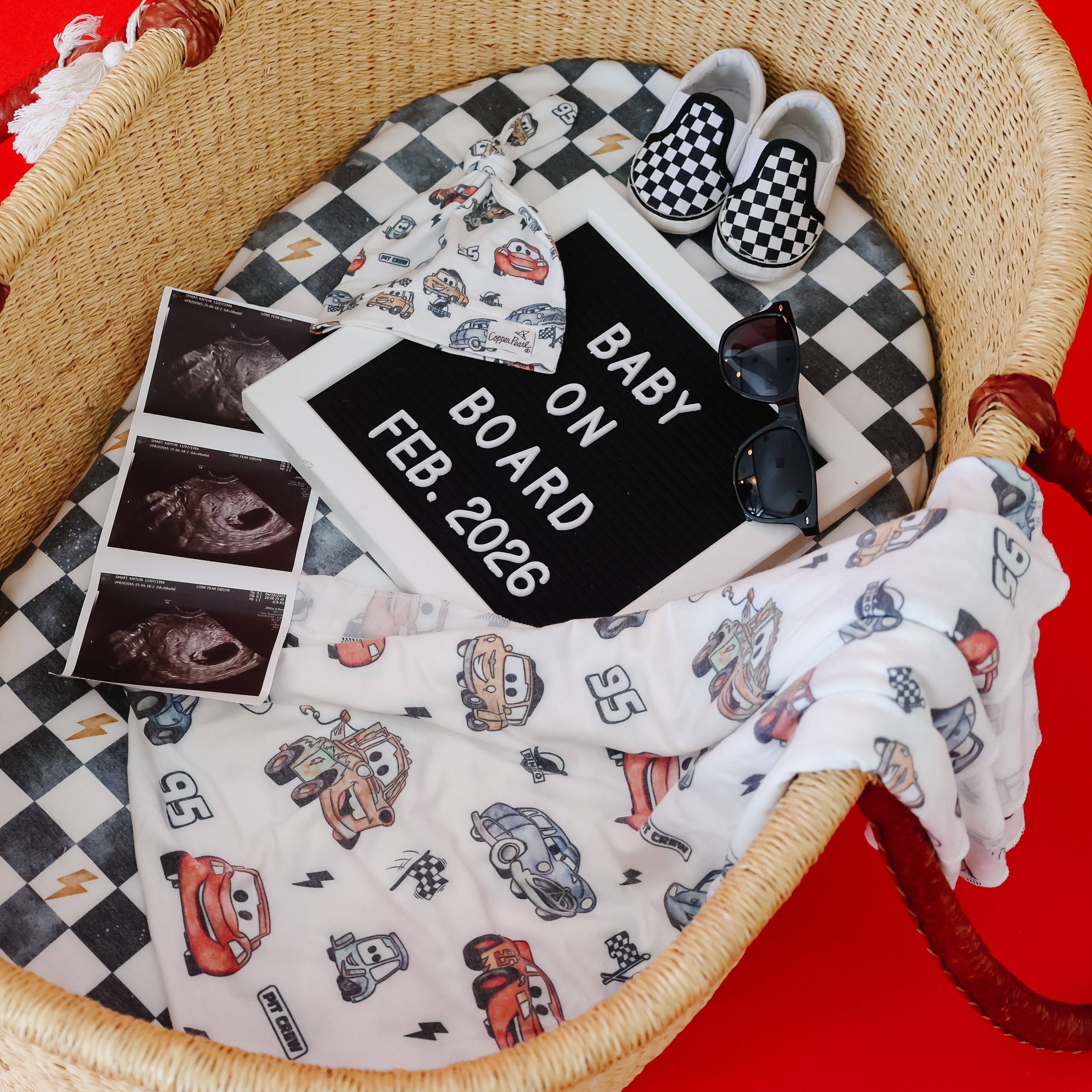 嬰兒提籃內擺放 Copper Pearl 汽車總動員麥坤款包巾、嬰兒帽、超音波照與迎接寶寶的英文字母板。