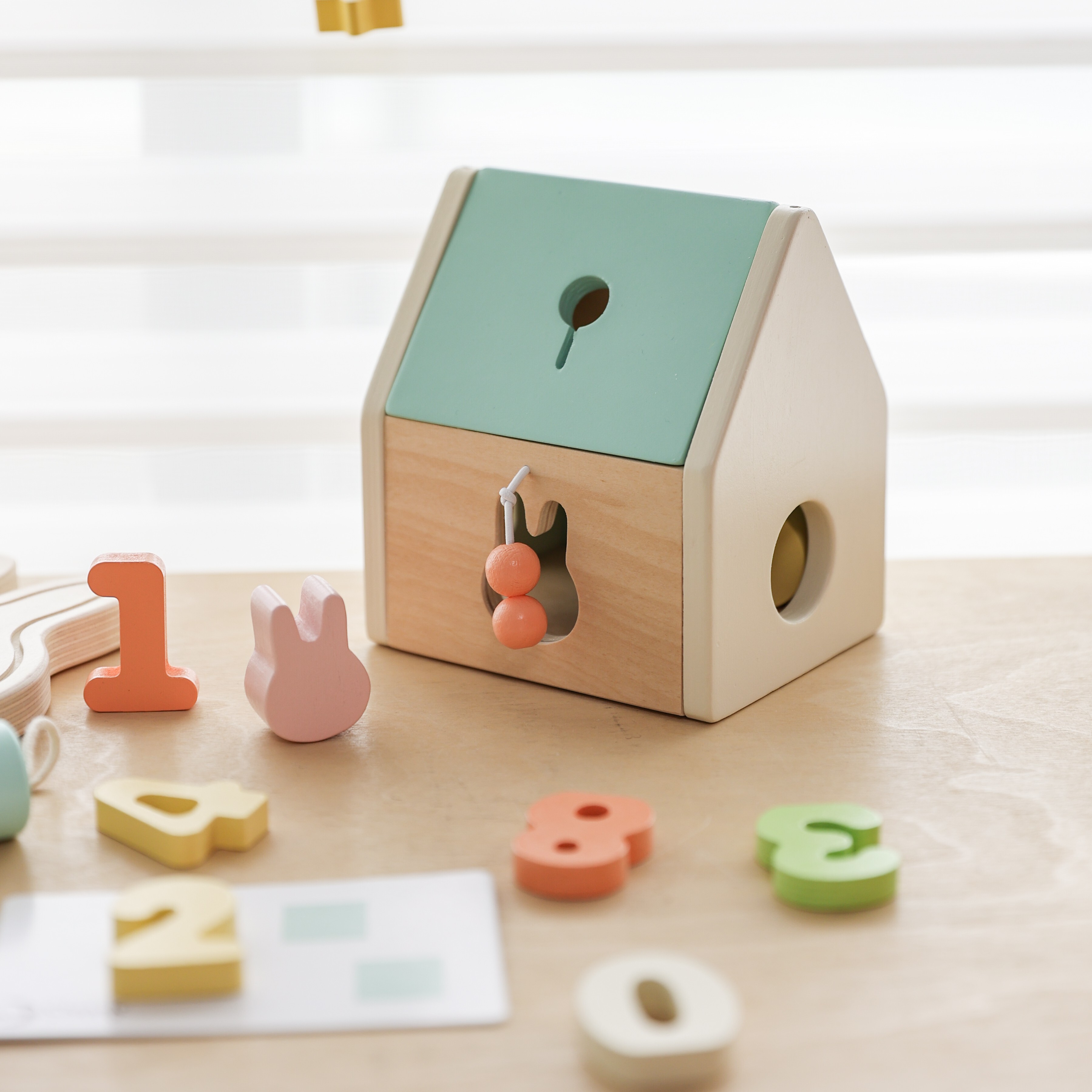木質桌面上擺放著 Classic World 幼兒認知啟蒙禮盒的造型小木屋，周圍散落著粉、橘、黃、綠等柔和馬卡龍色系的木製數字積木與粉紅色小兔子積木。