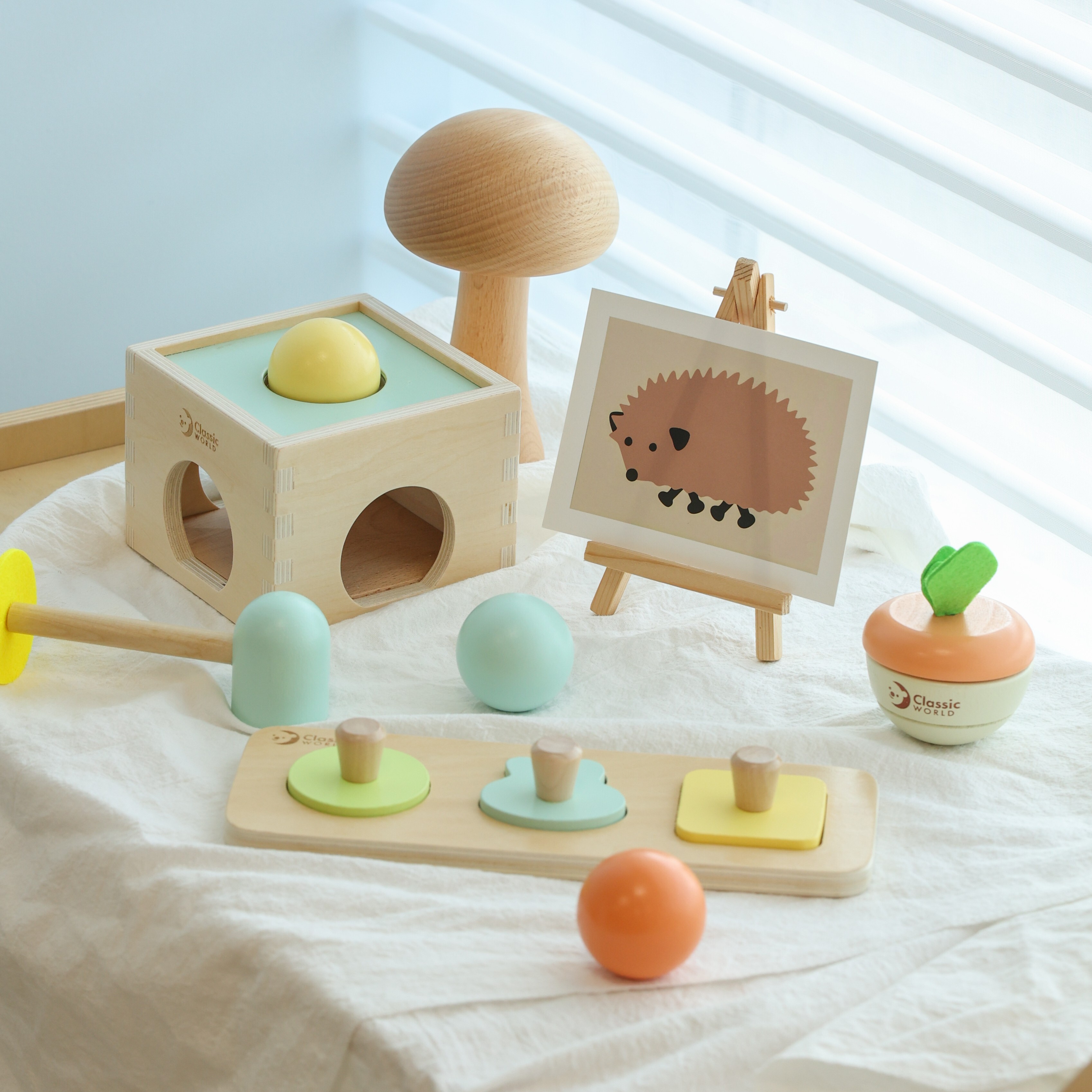 白色布面上擺放著 Classic World 寶寶手眼訓練禮盒的木製玩具，包含帶有黃色木球的敲擊台與木槌、幾何形狀抓握板、彩色木球，旁邊還點綴著刺蝟圖案的小畫架與木製蘑菇擺飾。