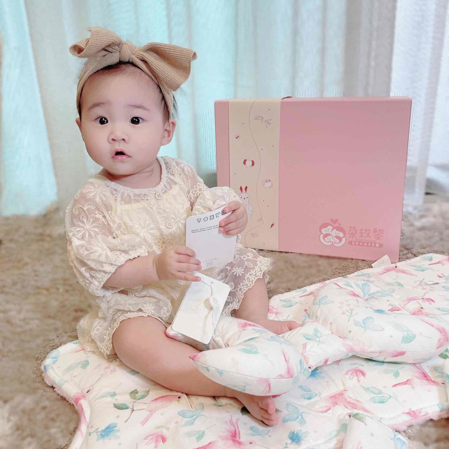 女寶寶坐在Maylily防晃枕與推車墊旁，後方為朶玫黎Tomali質感粉色彌月禮盒。 （Maylily推車配件3件禮盒組圖片提供：@kikibakikiby）