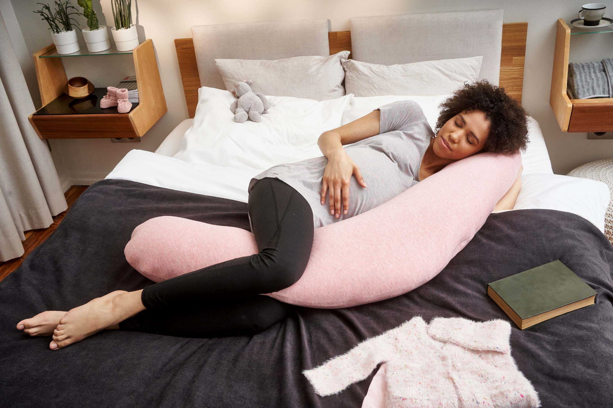 一張鳥瞰圖，顯示一位穿著灰色T恤和黑色打底褲的非裔孕婦在現代臥室的大床上閉眼休息。她側躺著，身體和腿靠在一個長條形、實心粉色的U型孕婦枕上，一隻手放在孕肚上。床頭櫃上有植物和嬰兒小鞋。