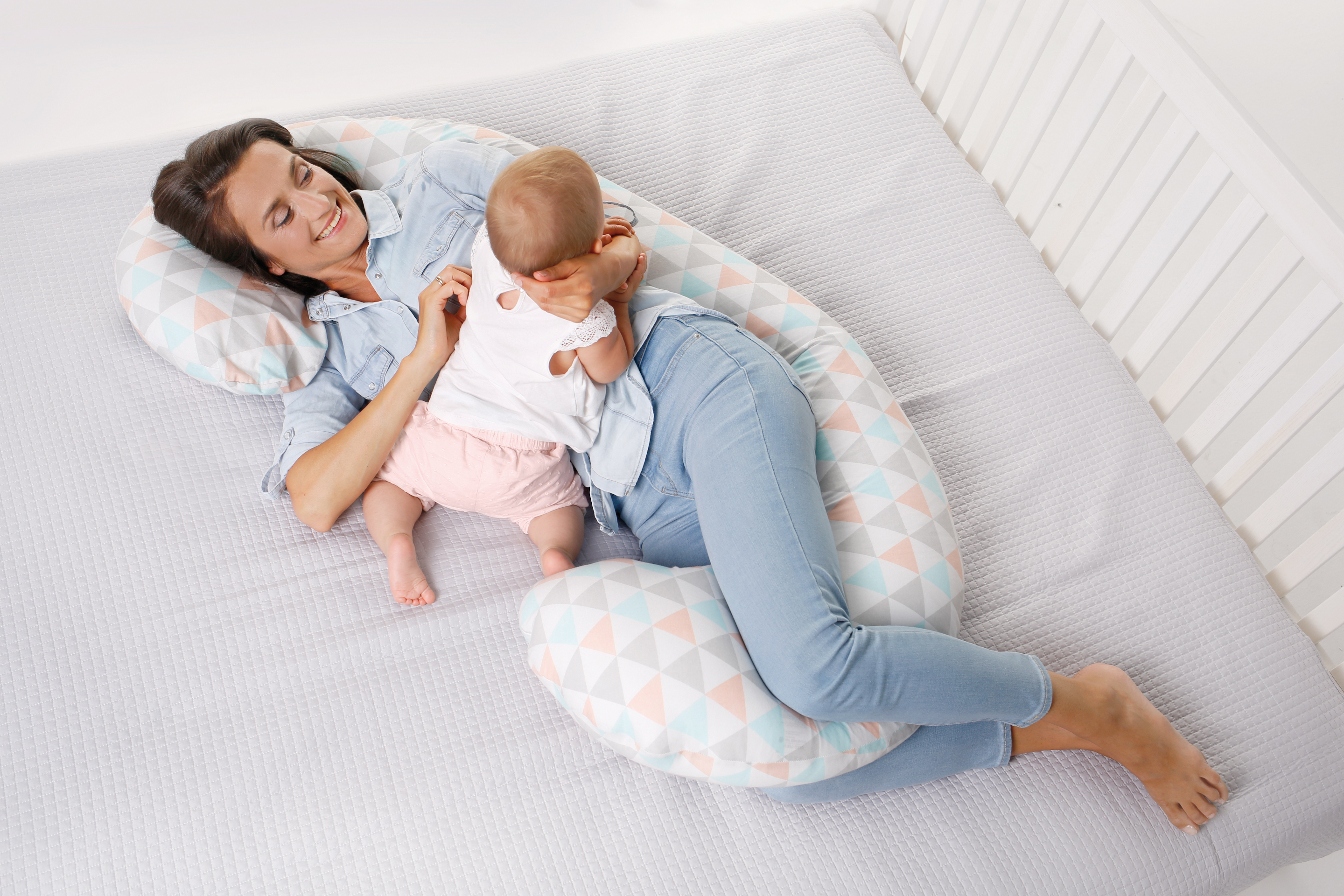 一張鳥瞰圖，顯示一位穿著牛仔衣的母親在鋪著灰色床單的白色嬰兒床內，躺在一個長條形、幾何三角形圖案的孕婦/哺乳枕上與她的寶寶互動，寶寶背對著鏡頭。
