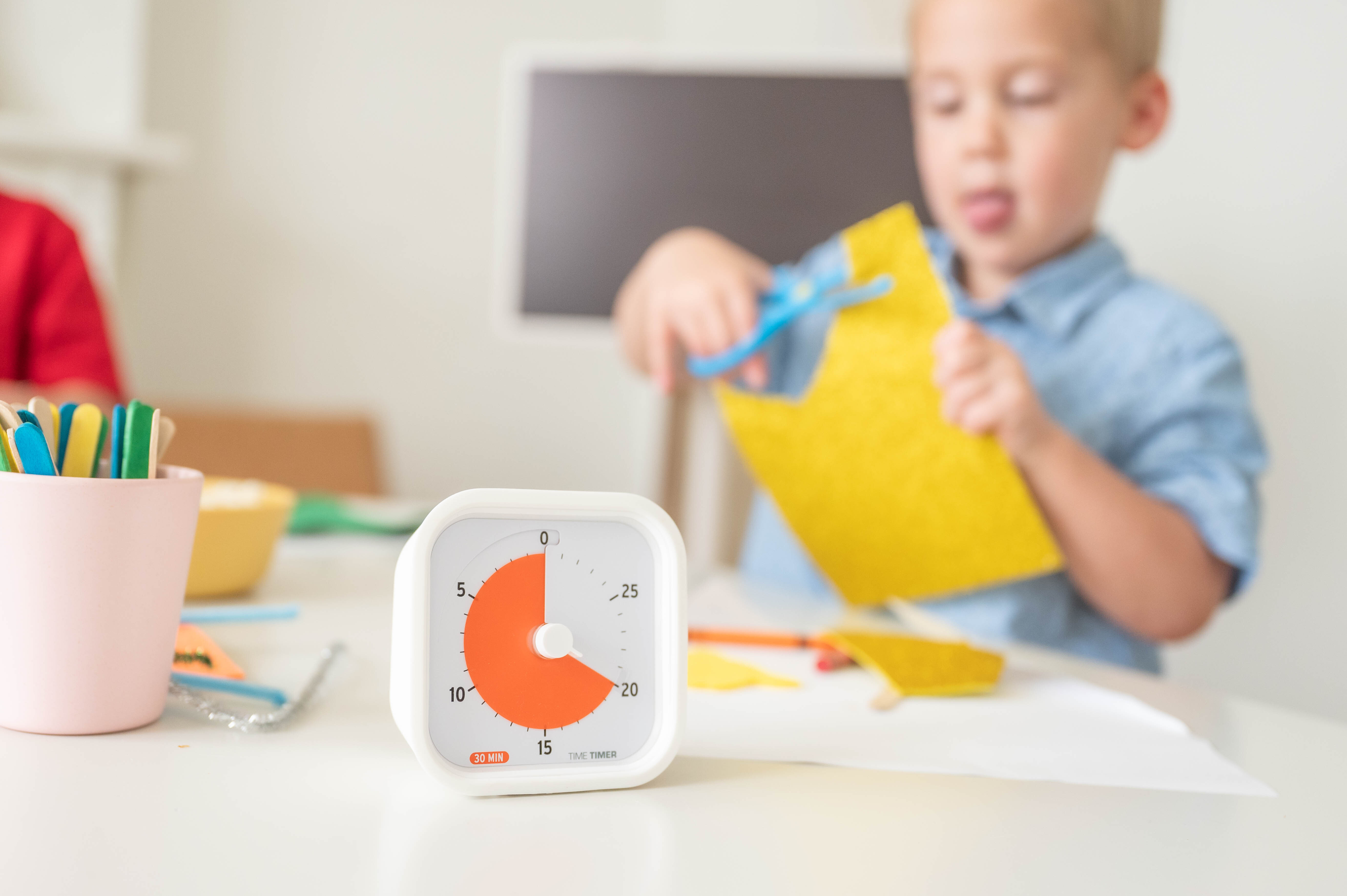 放在桌上的 Time Timer 視覺計時器，顯示剩餘橙色時間。背景模糊，一個男孩在剪紙。