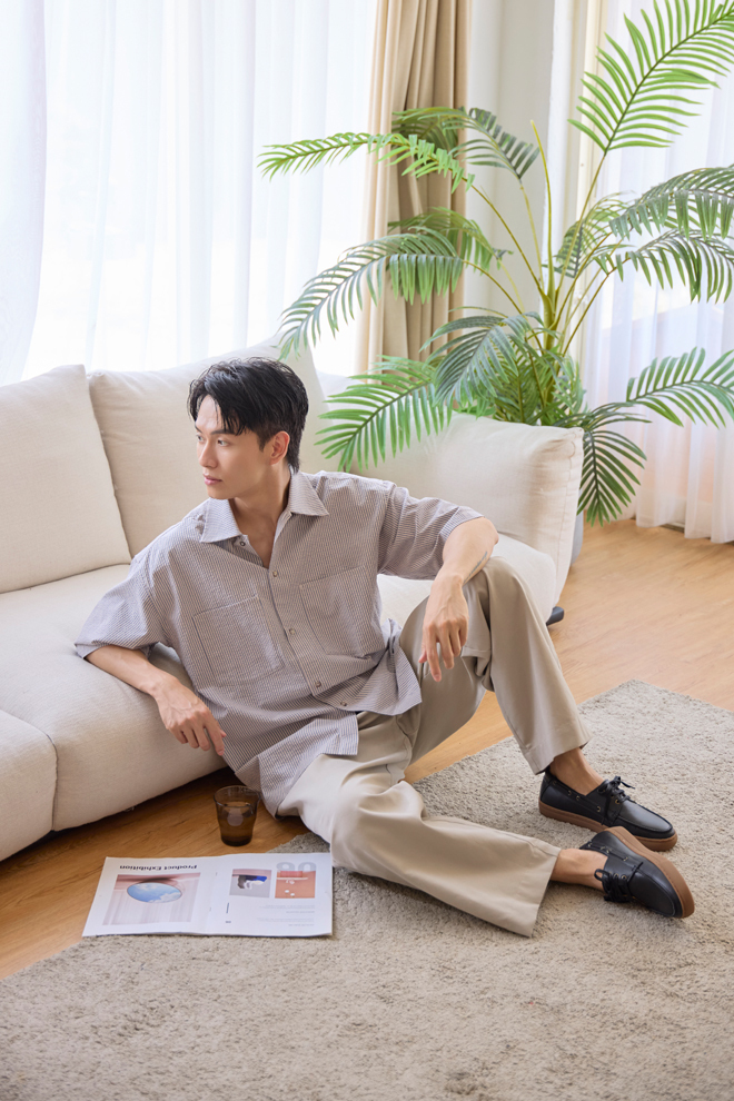 男士坐在沙發前的地毯上,穿著米色寬褲與林果黑色帆船鞋的生活情境照