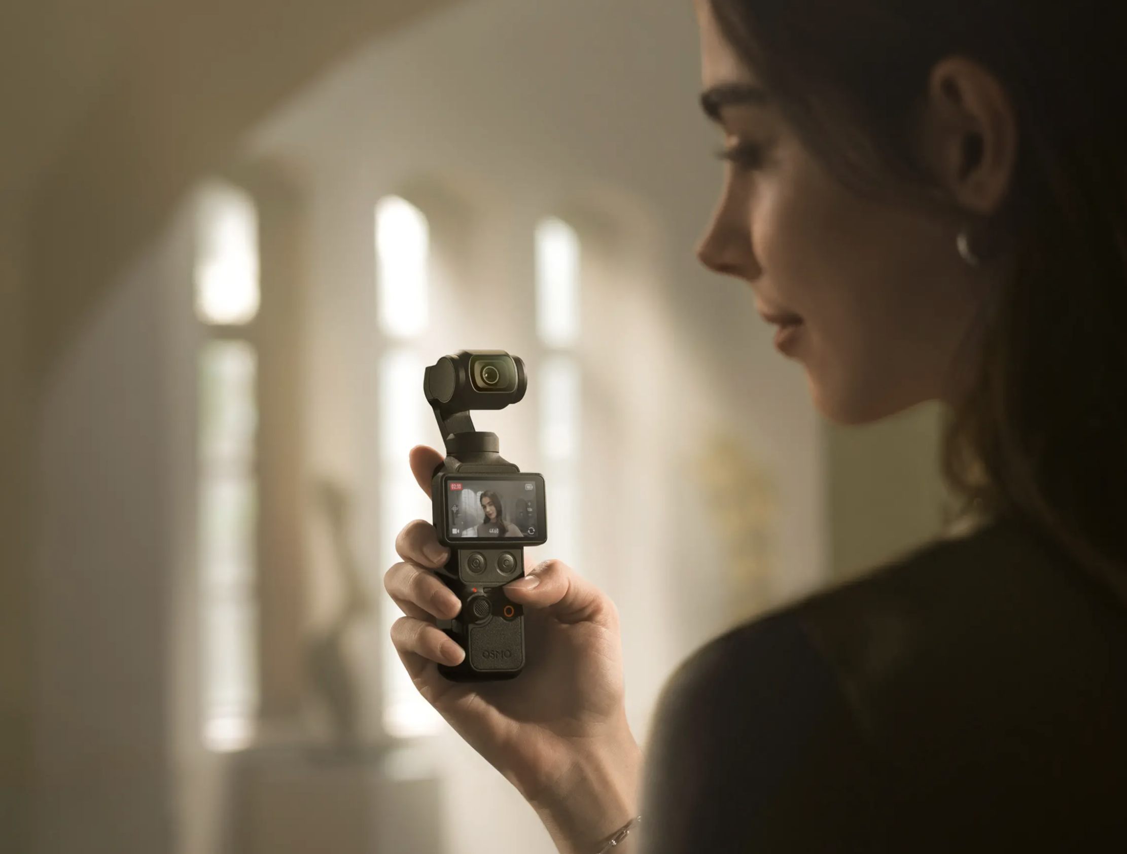 DJI Osmo Pocket 4 官方使用情境圖,女性側臉手持相機自拍,螢幕顯示 4K60 錄影中畫面,背景為柔和自然光走廊,呈現室內人像自拍的手持穩定攝影應用。
