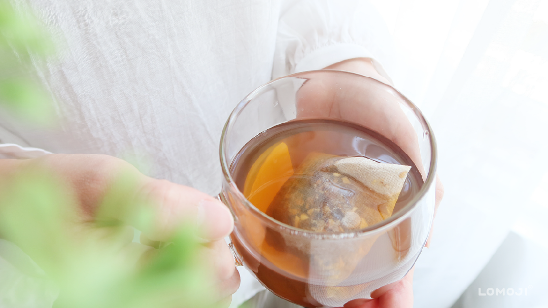 解決天氣熱想喝冰飲的替代方案:女子雙手捧著透明杯裝的琥珀色溫熱漢方茶,呈現夏季透過溫熱草本茶與居家按摩保養的放鬆日常