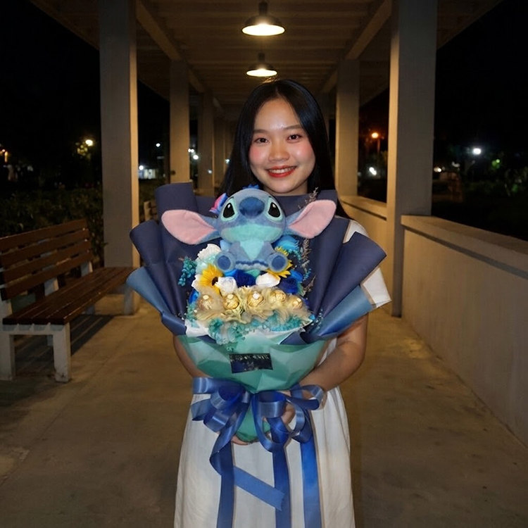 女生在夜間走廊手持史迪奇玩偶向日葵花束，搭配藍白玫瑰、金色巧克力與深藍色包裝緞帶