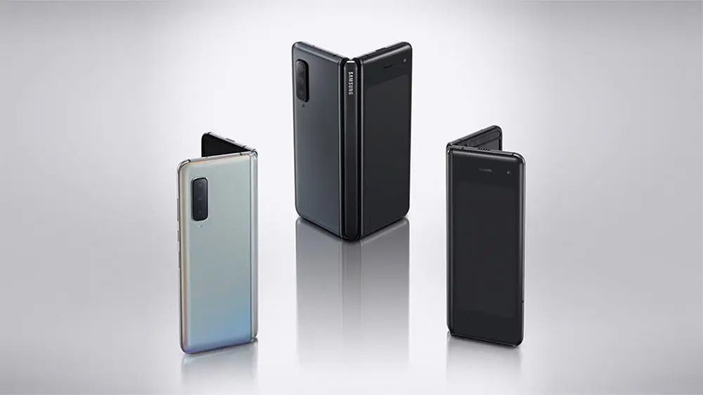 三星 Galaxy Z Fold 系列摺疊手機歷代機型陣列官方形象圖,三支手機皆呈半開摺頁狀立於灰白漸層反光地面,由左至右為銀色、深灰、黑色三種配色,展示機身摺疊角度與內外雙螢幕設計,背景為漸層灰白棚拍。