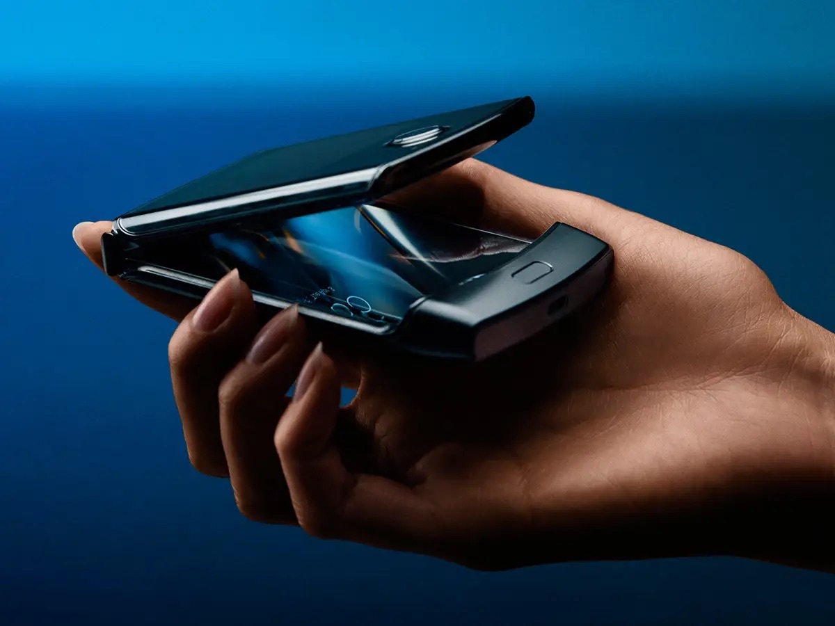 Motorola Razr 摺疊手機官方形象圖,手掌托著半掀開狀態的機身特寫,可見鉸鏈轉軸與內螢幕微亮畫面,機頂圓形相機模組清晰可見,背景為深藍色漸層。