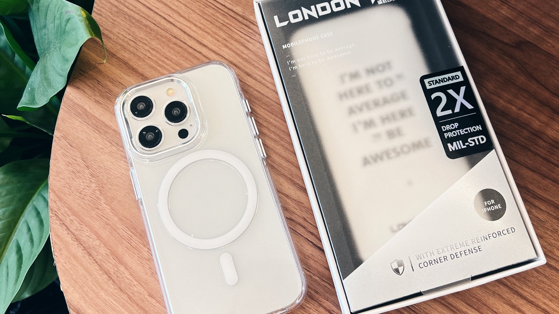 透明明岳殼 iPhone 與 LONDONIMG 包裝盒並放於木質圓桌，左側有綠色植栽，殼身通透可見機身原色
