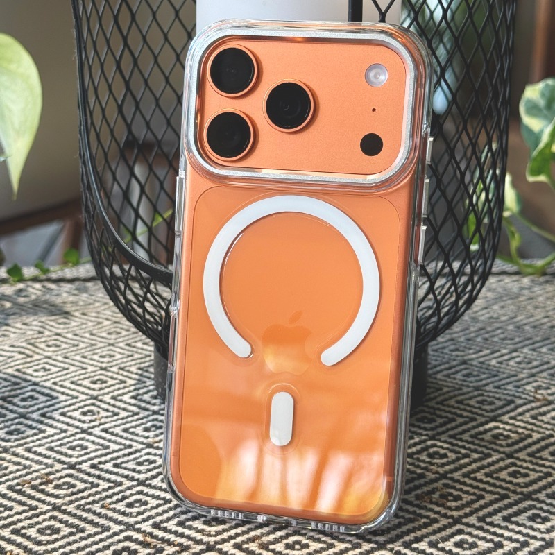 透明明岳殼包覆的橘色 iPhone 立於戶外金屬網籃前，背面可見白色 MagSafe 磁環