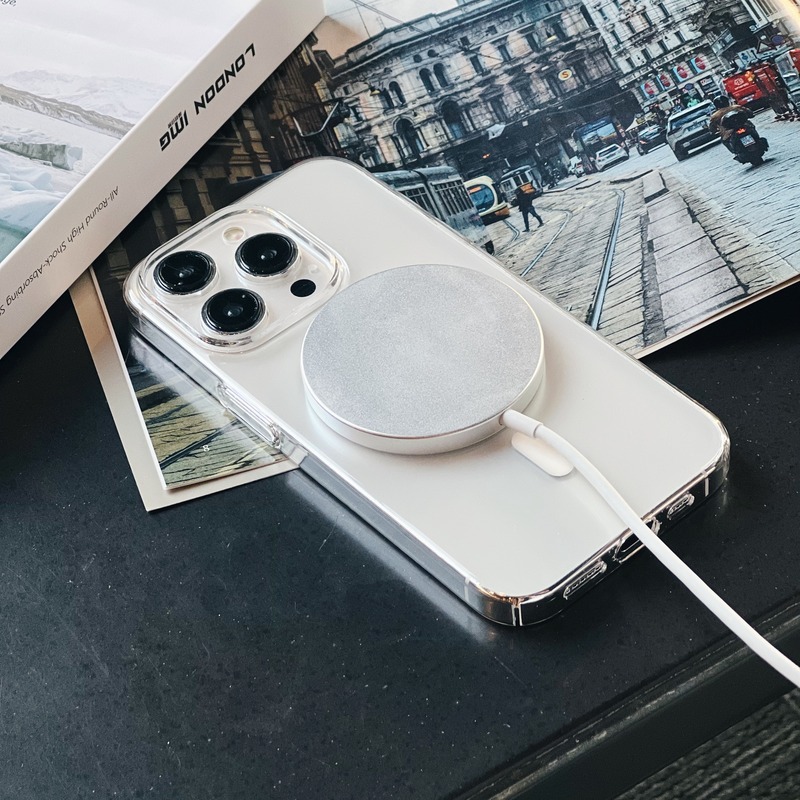 桌面上的透明極薄殼 iPhone，背面吸附白色 MagSafe 磁吸充電盤並連著充電線，旁有雜誌