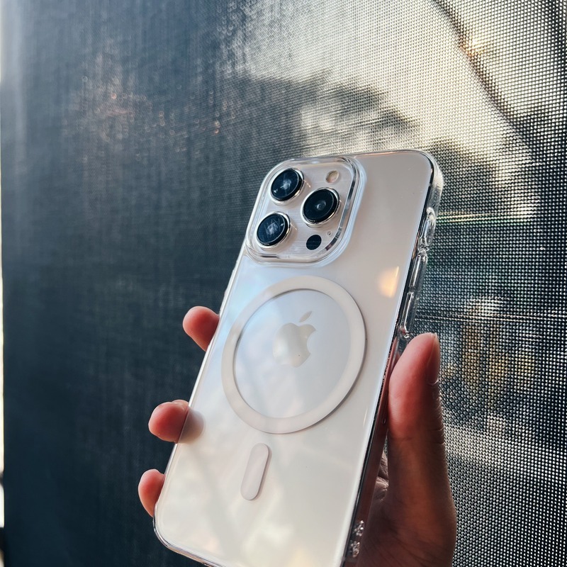 手持透明極薄殼 iPhone，鏡頭開孔與側邊按鍵開孔清晰可見，背景為紗窗與樹影