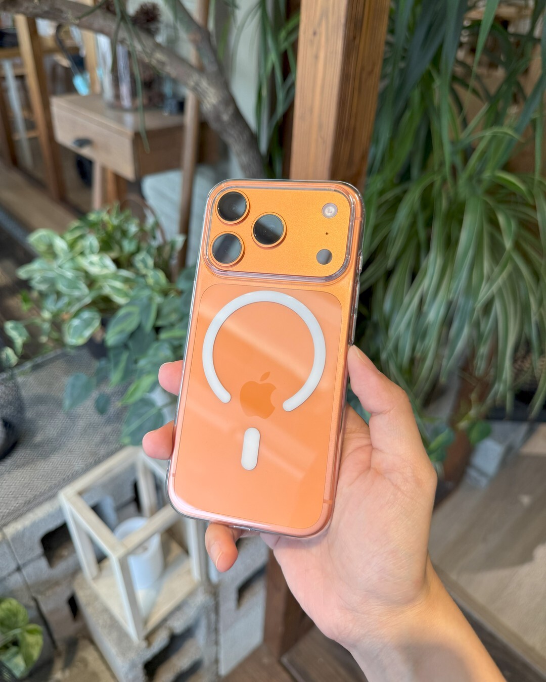手持橘色零手感 Lite Case 的 iPhone，背面 MagSafe 磁環清晰，背景為戶外植栽與木架