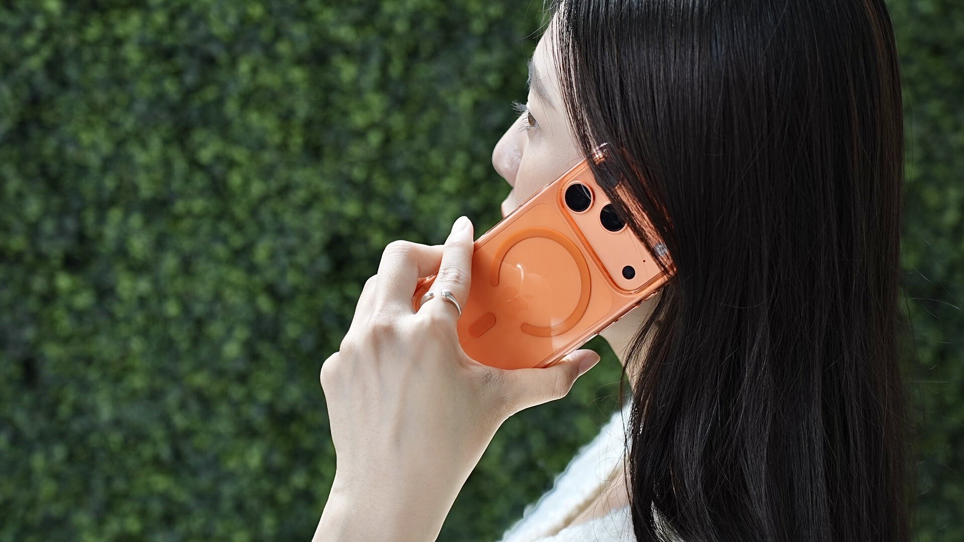 女性手持橘色零手感 Lite Case 的 iPhone 講電話，背景為戶外綠籬