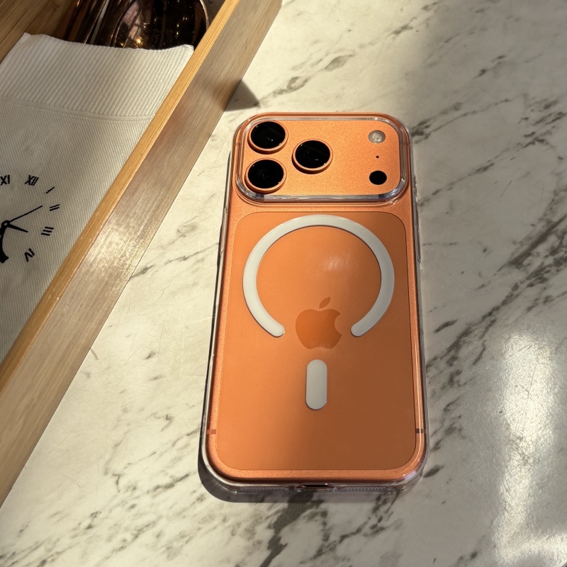 橘色零手感 Lite Case 包覆的 iPhone 平放大理石桌面，背面 MagSafe 磁環與鏡頭開孔清晰，左側為木質托盤