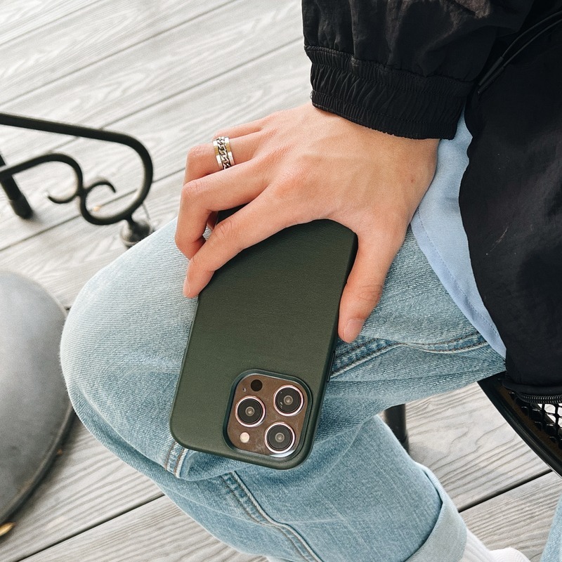 手持墨綠色 YU 皮革 MagSafe 磁吸殼的 iPhone，皮革面全包覆機身，四角與鏡頭框完整包住