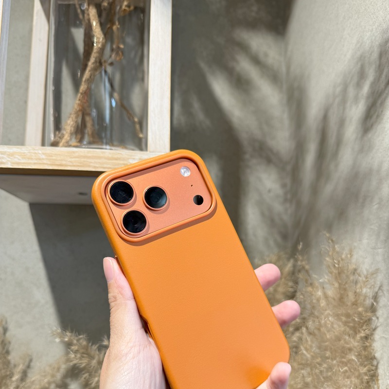 手持黃棕色皮革磁吸殼的 iPhone，皮革紋理在日光下呈現溫潤質感，背景為戶外芒草