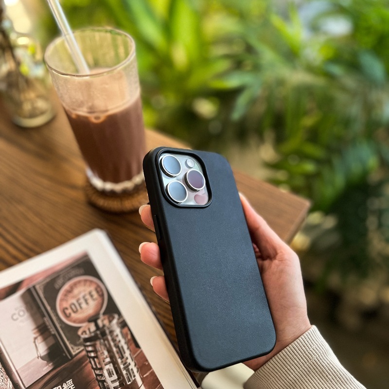 手持深藍色皮革磁吸殼的 iPhone 於咖啡與雜誌前，皮革表面乾淨無指紋痕跡