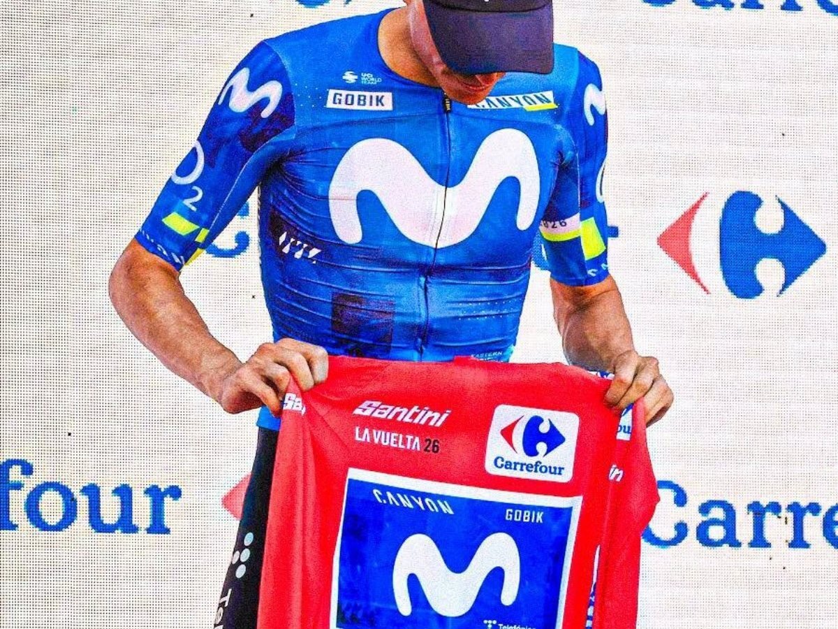 Enric Mas 在 2026 環西第 8 站頒獎台上身穿 Movistar 車隊青藍色車衣，雙手手持紅衫向傷退的 Tadej Pogačar 致敬