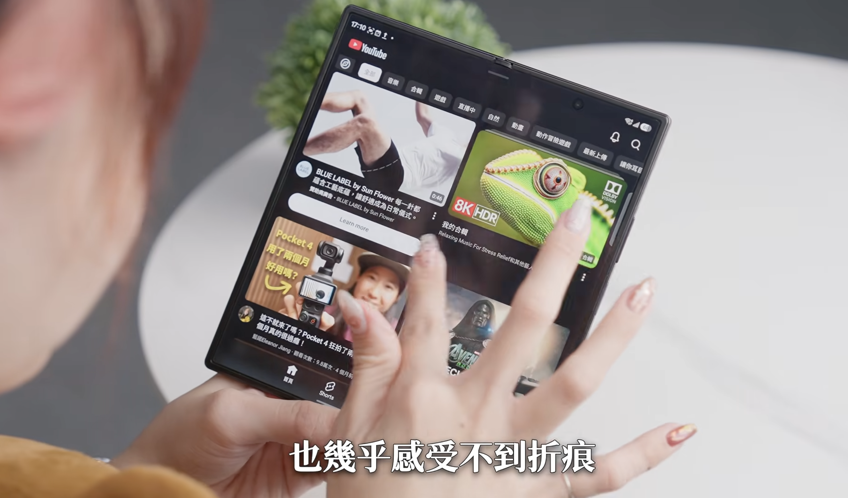手指滑過摺疊手機展開後內螢幕中央摺痕處的評測影片截圖,螢幕顯示 YouTube 首頁推薦影片清單,畫面下方字幕為「也幾乎感受不到折痕」,用於展示摺疊螢幕摺痕在觸摸與視覺上的實際表現。