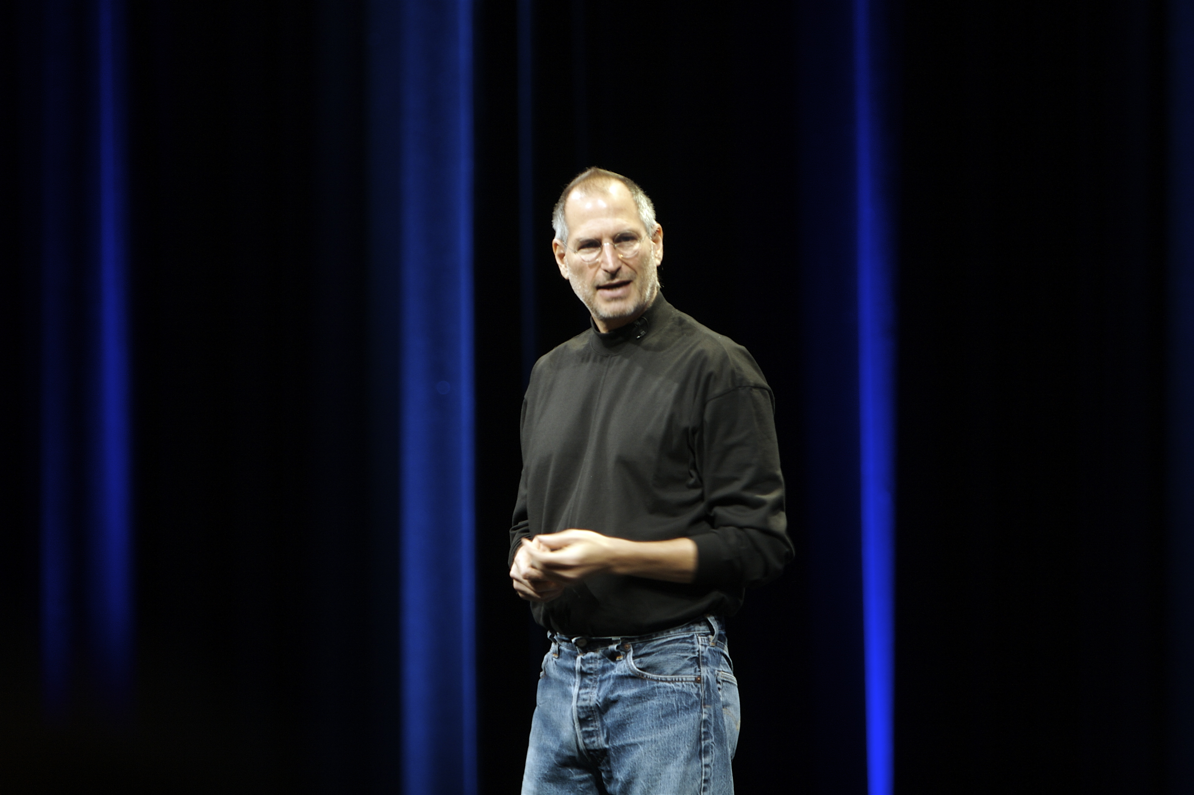 Steve Jobs 身穿黑色高領上衣與藍色牛仔褲,站在深藍色舞台布幕前雙手交握、面向觀眾發表演說的現場實拍,呈現 Apple 發表會經典的簡約穿著與舞台風格。