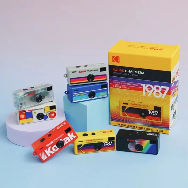Kodak Charmera 鑰匙圈數位相機官方商品圖,七款不同配色與圖案(透明、白底幾何圖形、灰底彩條、藍色、紅色「Kodak」大字、黃色「1987」復古款、黑色彩虹條紋)陳列於粉藍與淡紫色展示台,右側為黃色外包裝盒,標示「KODAK CHARMERA Keychain Digital Camera」、「Inspired by the Kodak Flino」、「7 Styles in Total」與「The Mini Camera & Blind Box All in One」。