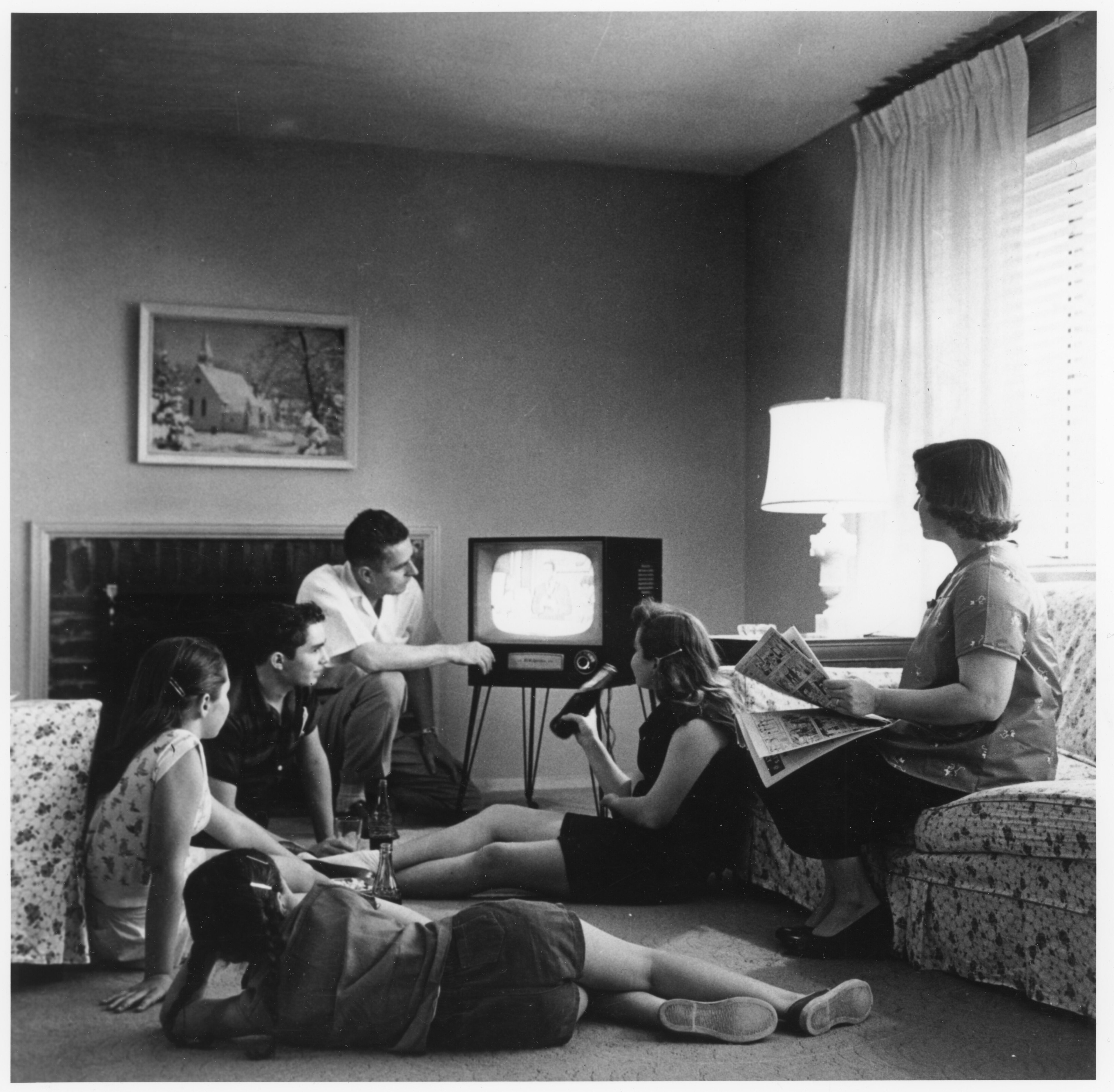 1950 年代美國家庭客廳圍坐觀看電視的黑白歷史照片,一家人或坐或趴於地毯與花布沙發上,視線集中於一台立在細腳座上的 4:3 近方形映像管電視,其中一人伸手調整電視,另一人手持報紙,背景為壁爐、教堂雪景畫作與透著光線的窗簾,呈現早期電視普及時的家庭觀影情境。