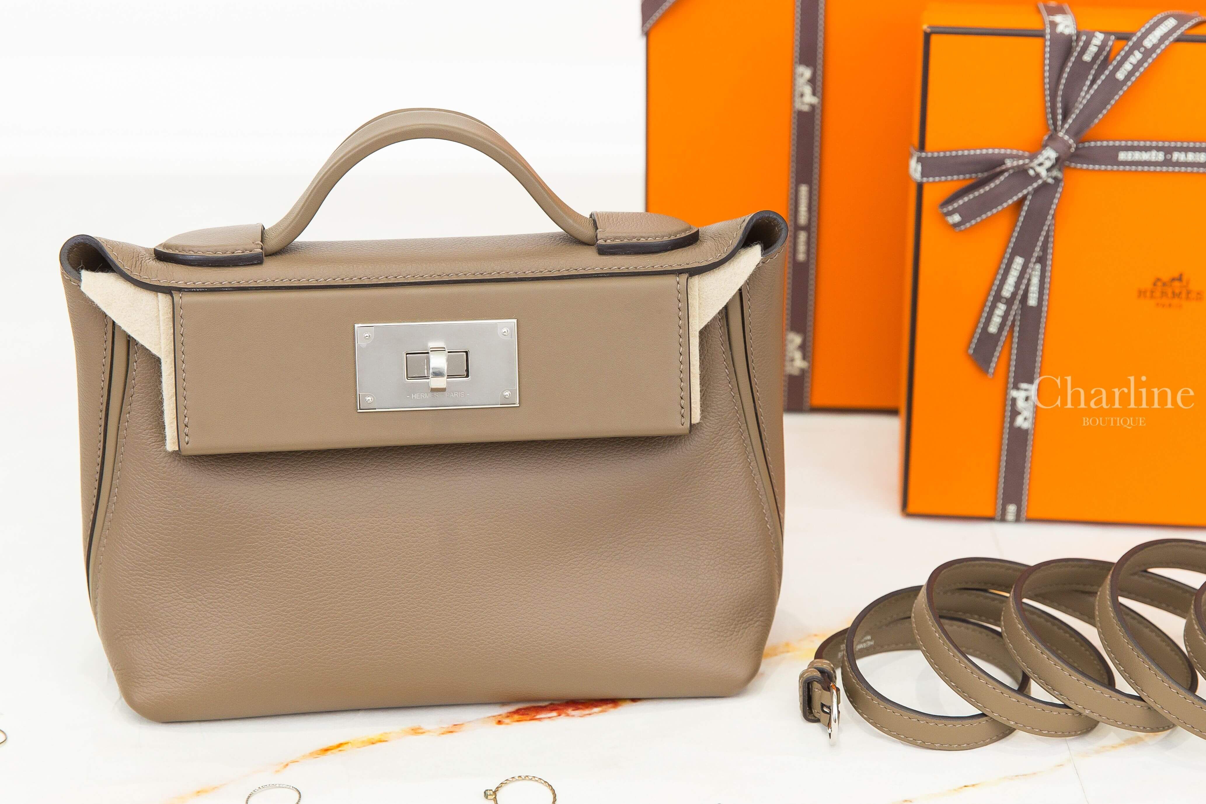 Hermès Mini Kelly 2 Etoupe 大象灰手提肩背包正面展示，搭配銀色旋轉鎖扣、弧形皮革提把與可拆式長肩帶，背景陳列愛馬仕橘色禮盒。