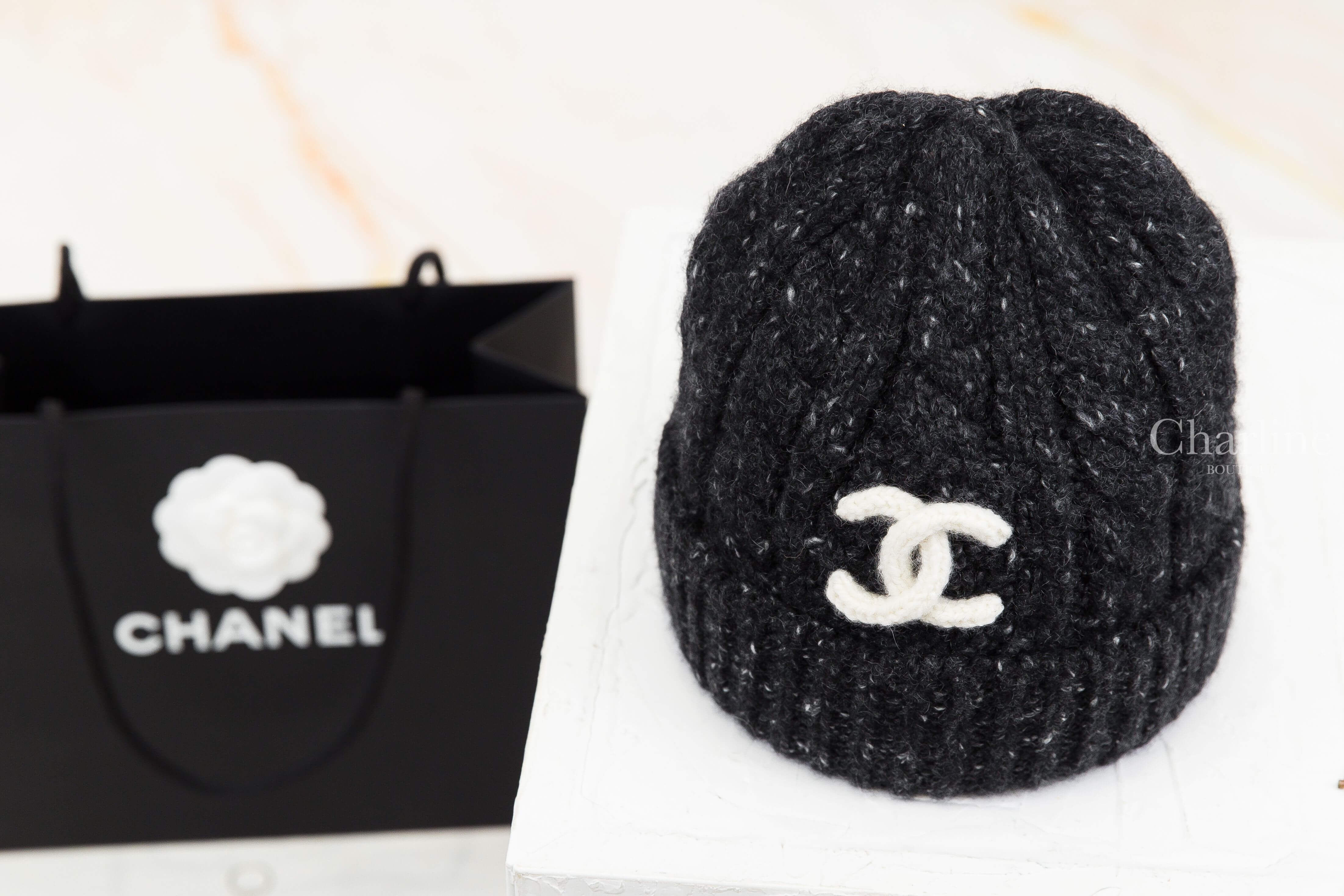 CHANEL 香奈兒黑色白雪花喀什米爾針織毛帽正面展示，採用立體羅紋針織設計，帽身點綴細緻白色混紗，正面搭配白色立體雙 C Logo，旁邊陳列 CHANEL 黑色品牌提袋。