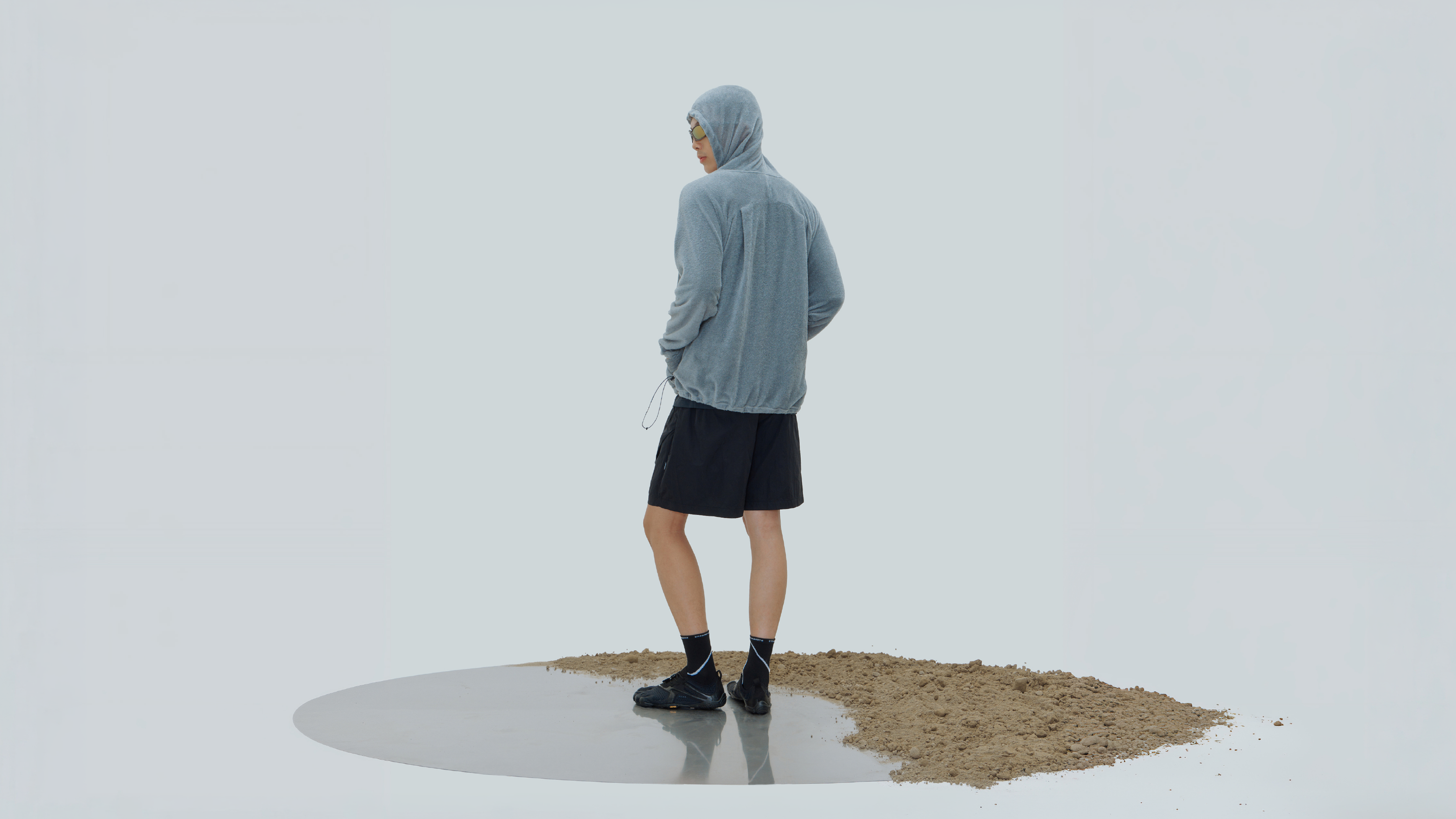 nozzle quiz® Octa® Ninja Hoodie 日常造型,模特兒穿著灰藍色連帽上衣與黑色短褲,以背側姿態站立於水面與土壤交界的攝影場景。