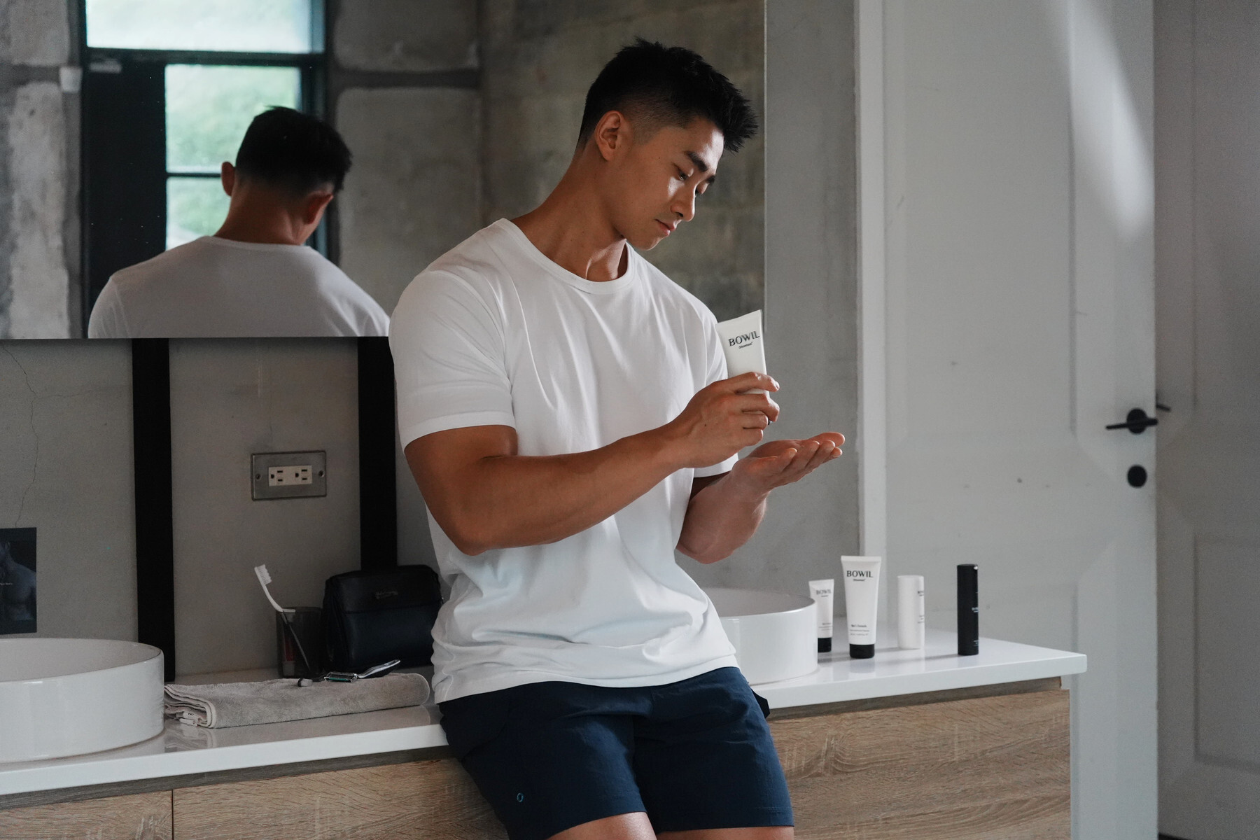 穿白色 T 恤的男子在洗手台前拿著 BOWIL 保養品,準備進行日常保養。